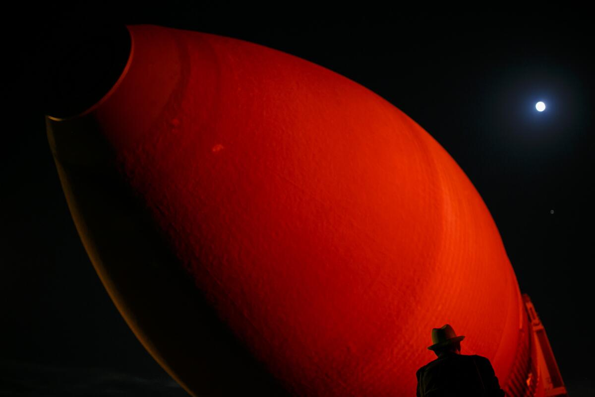 A man stands near a big orange space shuttle external fuel tank.