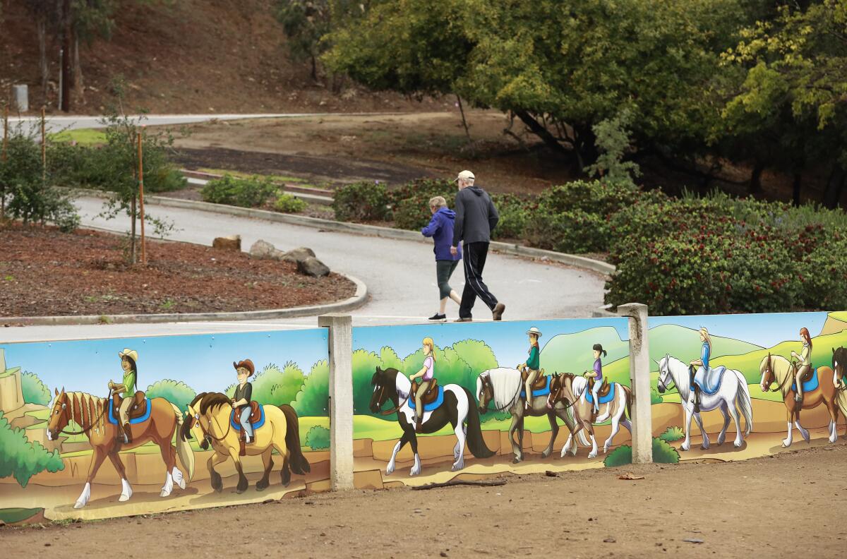 A mural depicting children riding ponies covers a fence behind which two people pass along a walkway