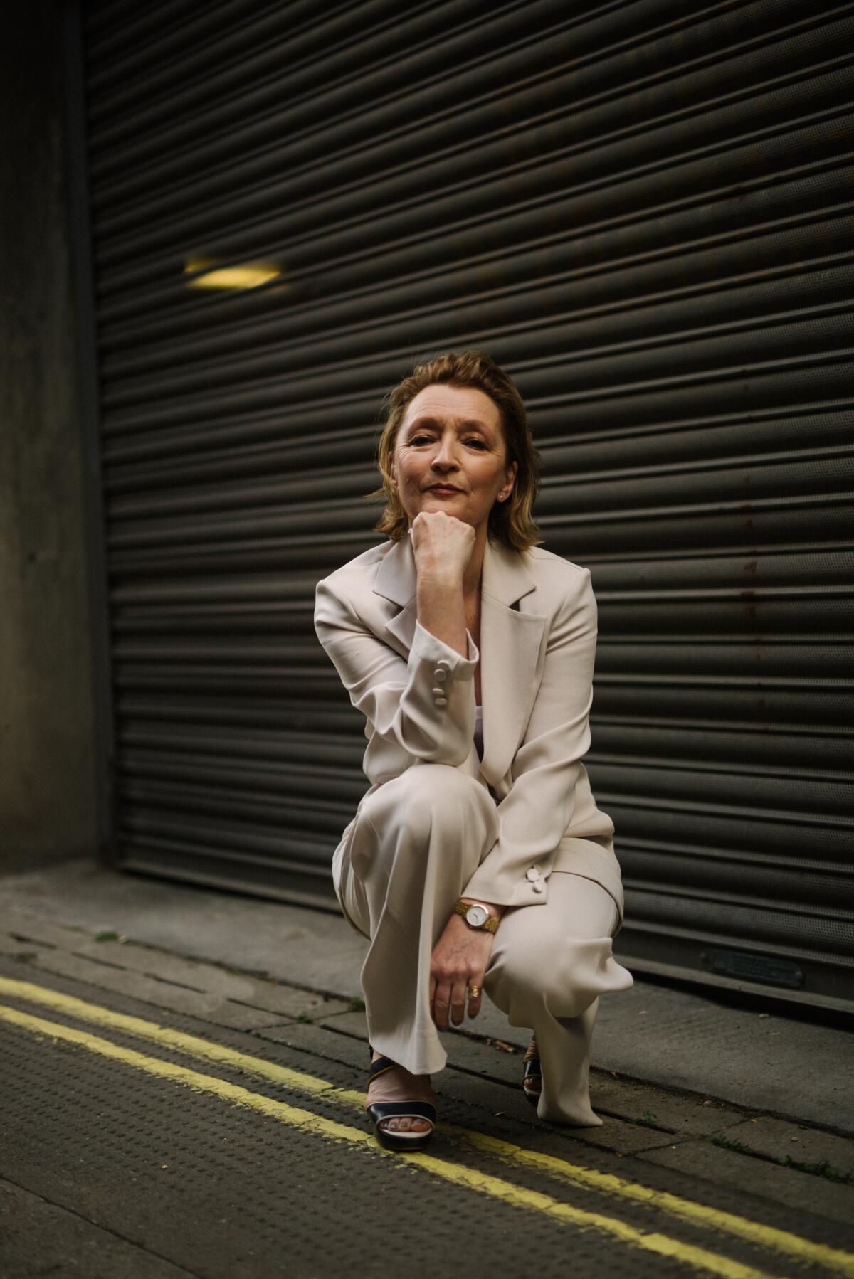 English actor Lesley Manville crouches down against an exterior wall, hand to her chin, for a portrait.