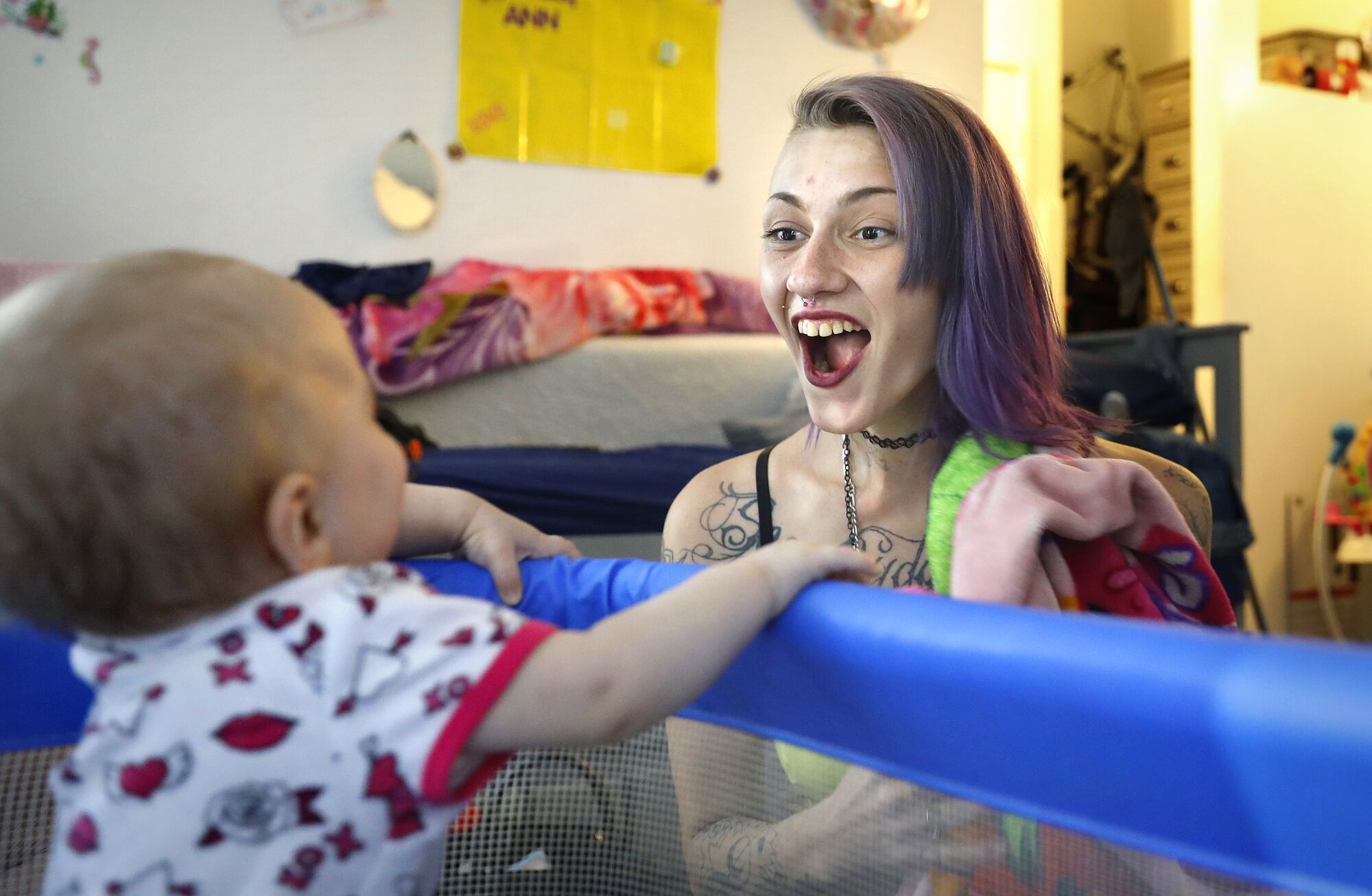 A woman smiling with an open mouth at a baby who is inside a playpen.