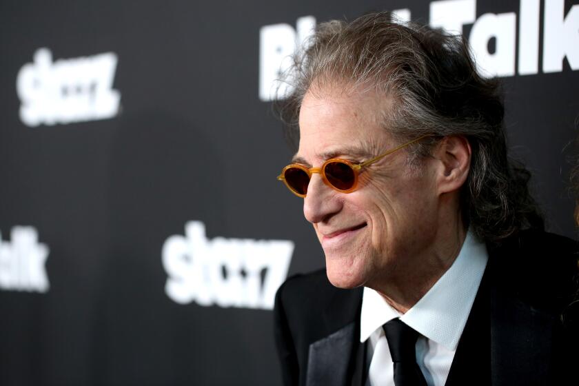 Richard Lewis wears yellow-rimmed sunglasses and smiles in a suit at a premiere 
