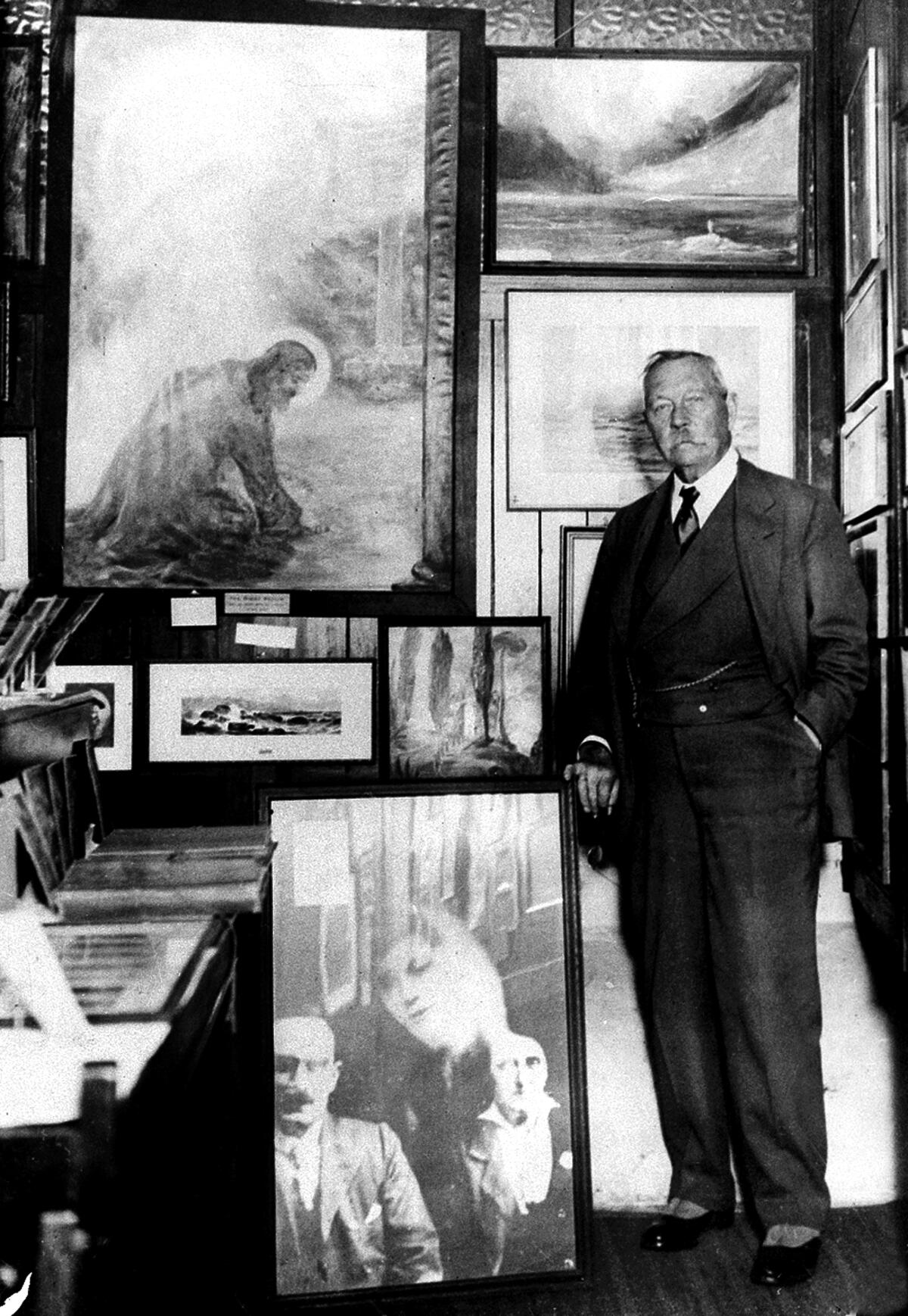 Arthur Conan Doyle, wearing a suit, stands beside a wall of paintings 