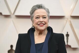 Kathy Bates in a black dress with a thick collar and wide shoulders smiling at the Oscars red carpet