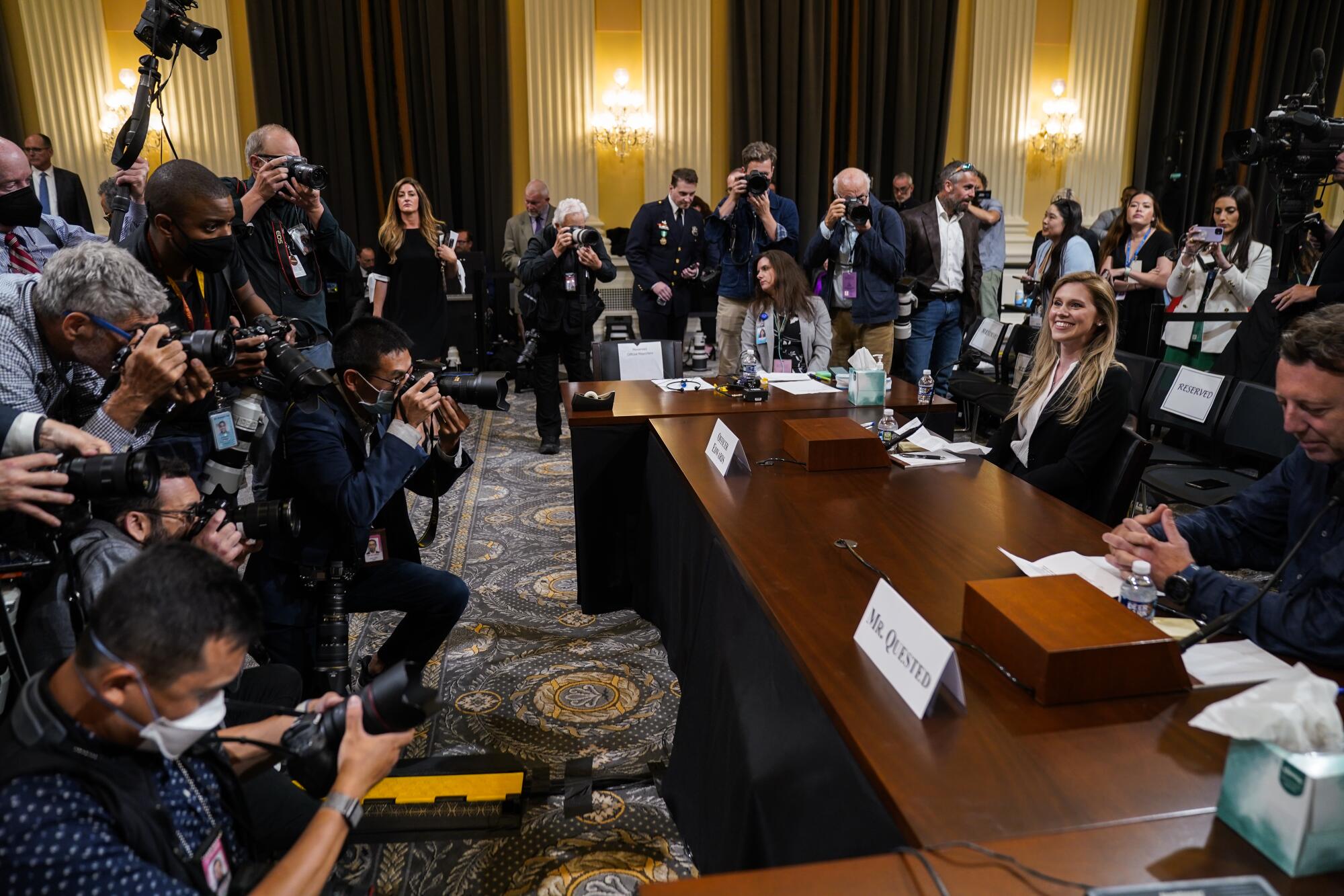 A bank of photographers taking pictures of a woman and a man seated at a witness table in an ornate hearing room