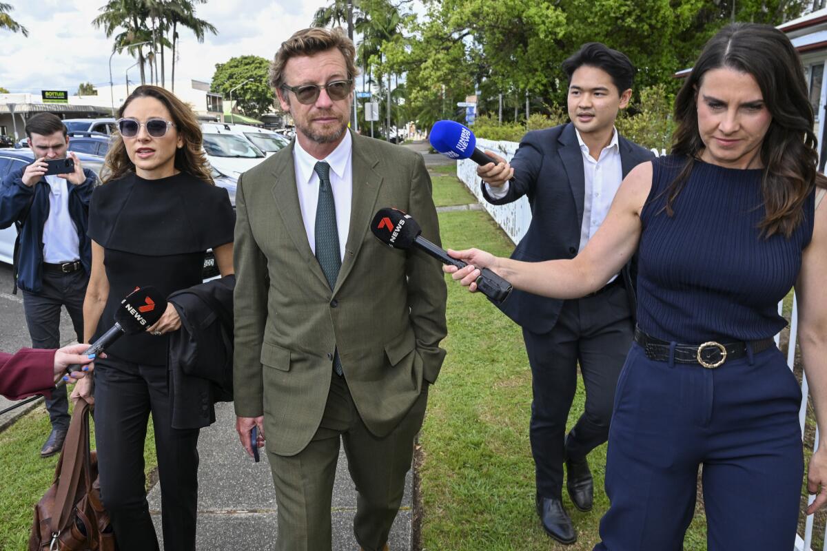 Simon Baker walking outdoors in an olive green suit surrounded by reporters thrusting microphones at him