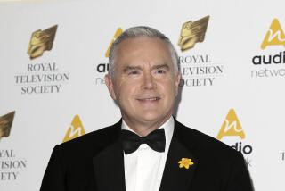 A man with short silver chair in a black tuxedo and a yellow flower broach poses against a white backdrop wiyh black text