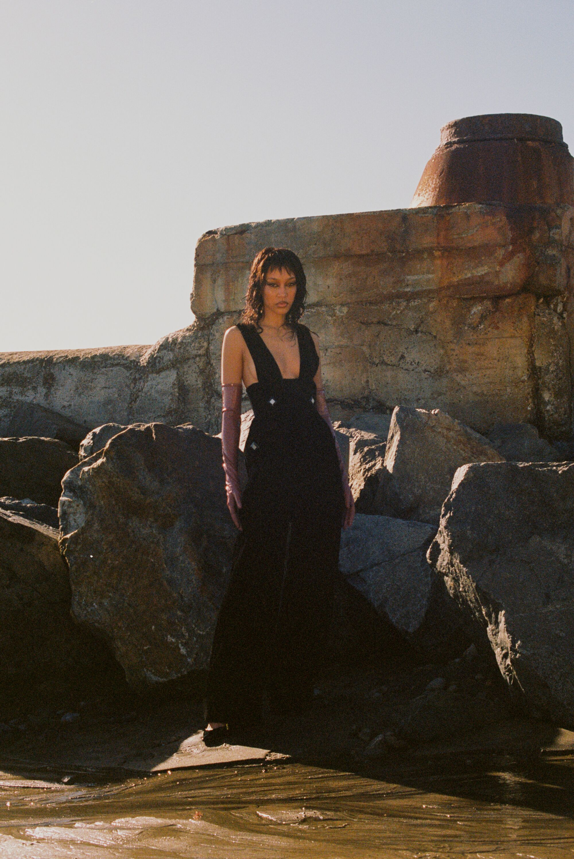 A model poses in an all-black dress on the beach, with rocks behind her.