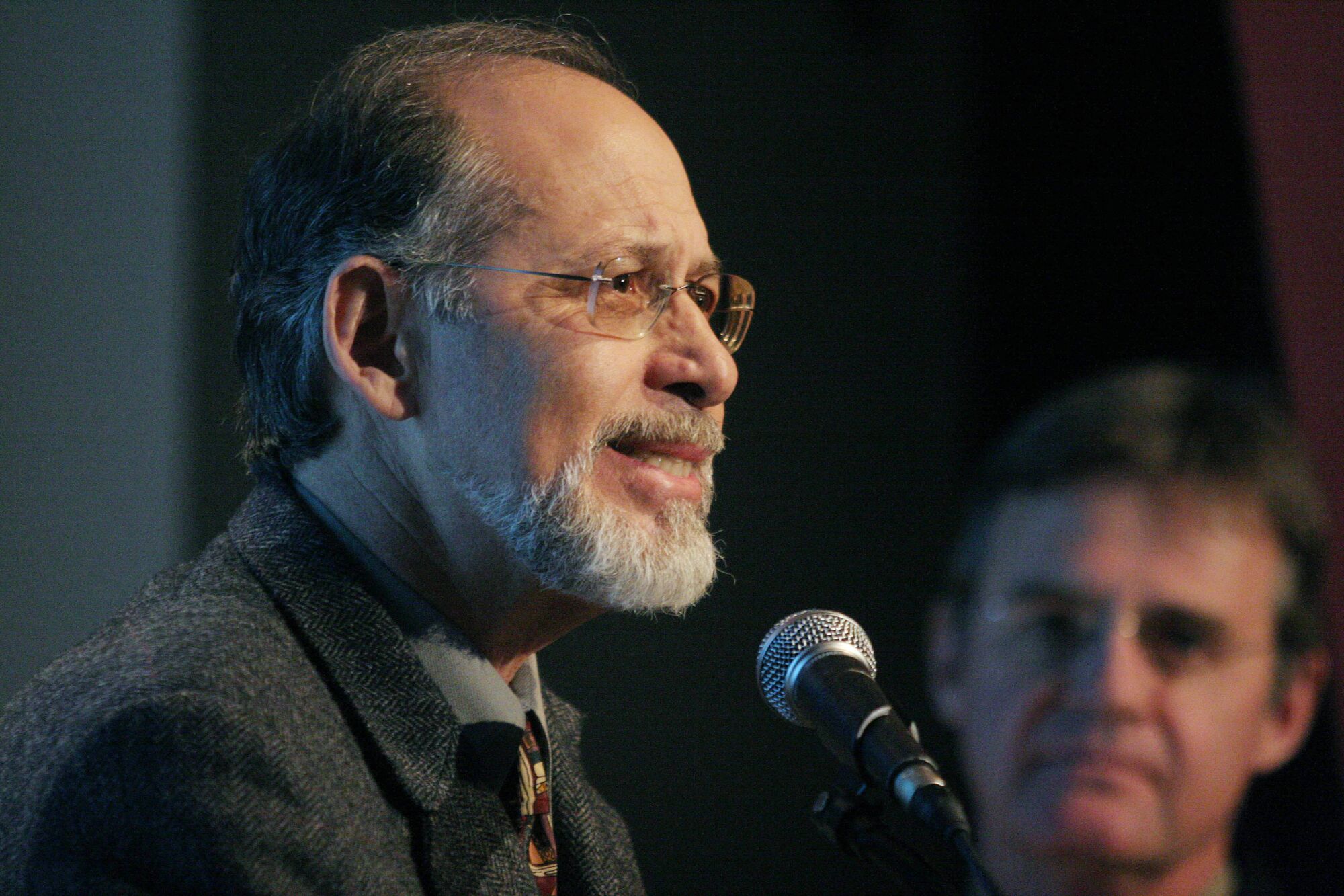 A man speaks into a microphone as another watches him in the background.