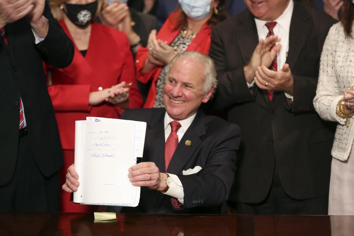 A seated man in a suit smiles and holds up papers as people clap behind him. 
