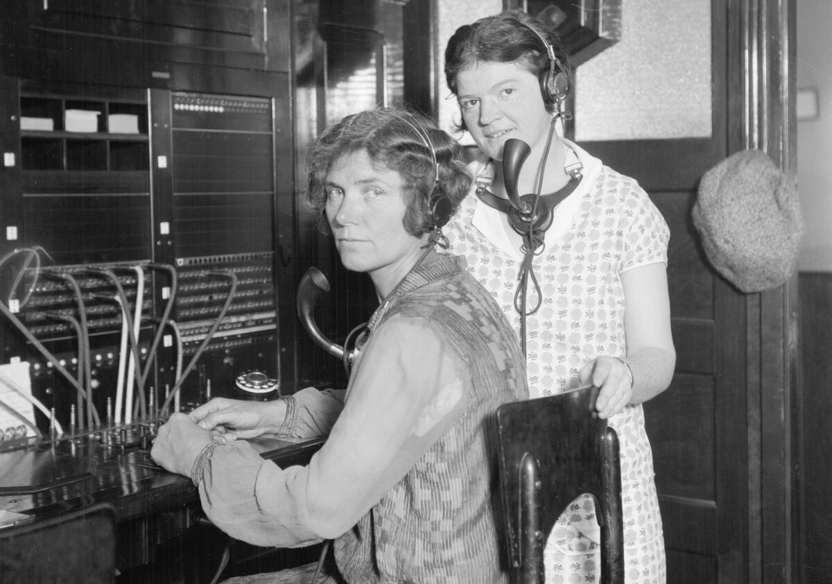 Two women in front of a telephone switchboard. Each wears headphones and a large mouthpiece.