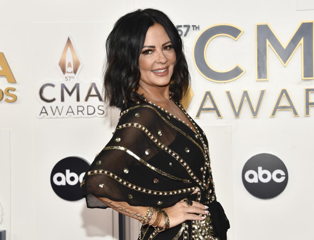 Sara Evans, in a black and gold gown, turns to the side with her hand on her hip in front of a CMA Awards backdrop