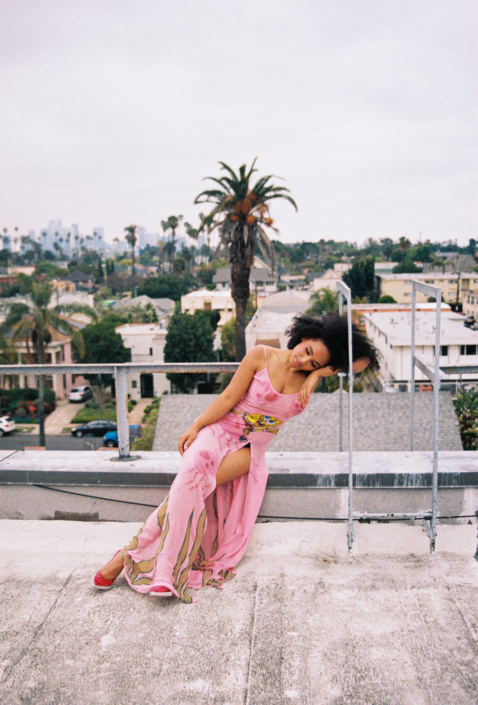 A woman wearing a long pink dress leans against the fire escape on a rooftop.
