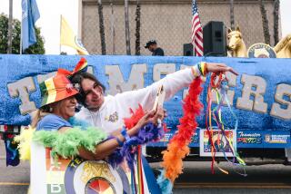 Two women dressed in rainbow outfits and accessories taking a selfie in front of a parade float on the street.