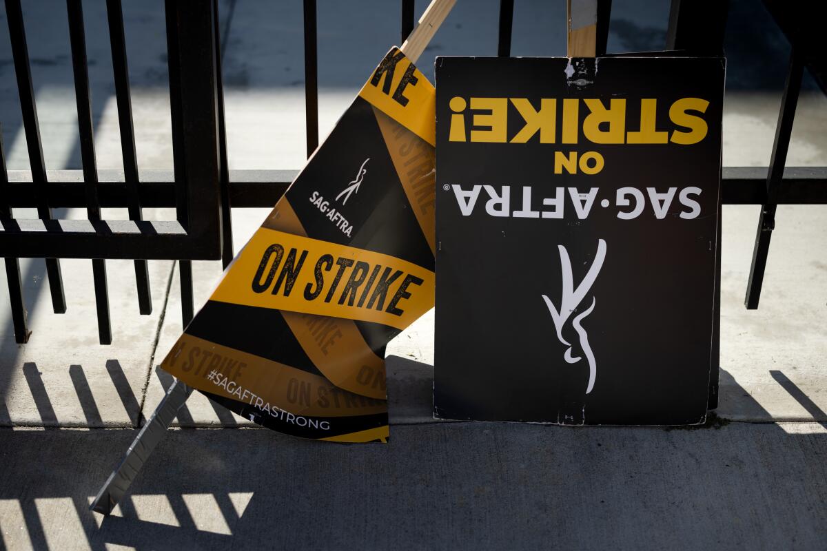 Black and yellow "SAG-AFTRA on strike" picket signs lie on the ground against a black metal fence
