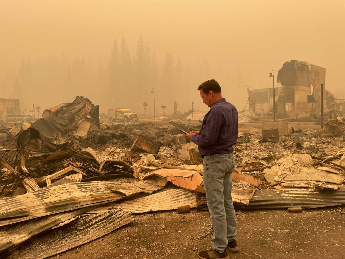 A man stands in front of the burned ruins of a home amid orange smoky haze
