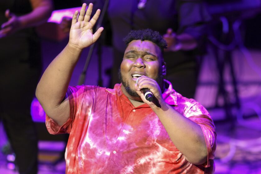 A man in a tie-dye shirt raising a hand into the air and singing into a microphone