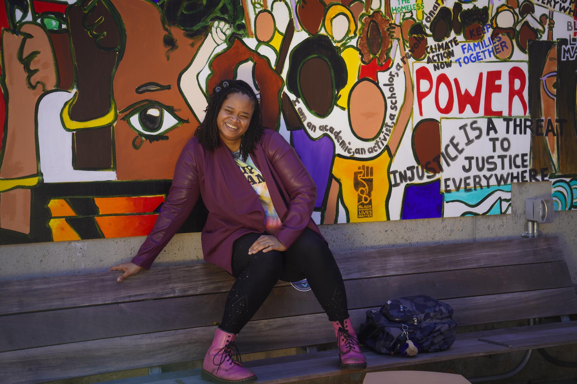 A woman sits on a bench next to a section of a colorful mural that displays the symbol for “Black Disabled Lives Matter.”