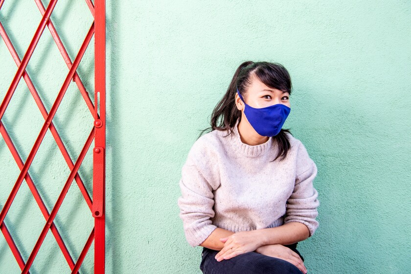 A woman in a ponytail and face mask sits for a photo against a green wall.