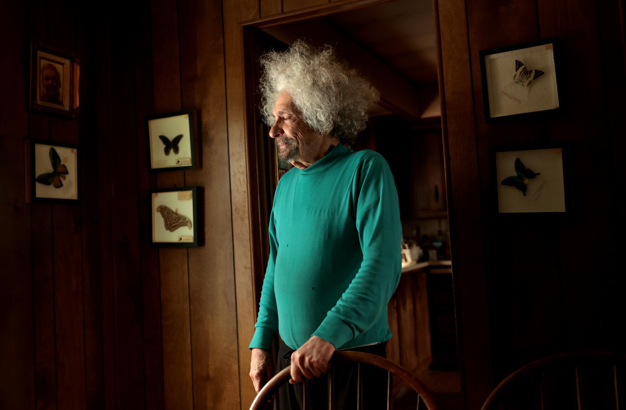 A man with bushy gray hair, in a turquoise-colored shirt, stands in a wood-paneled room with framed butterflies on the wall 