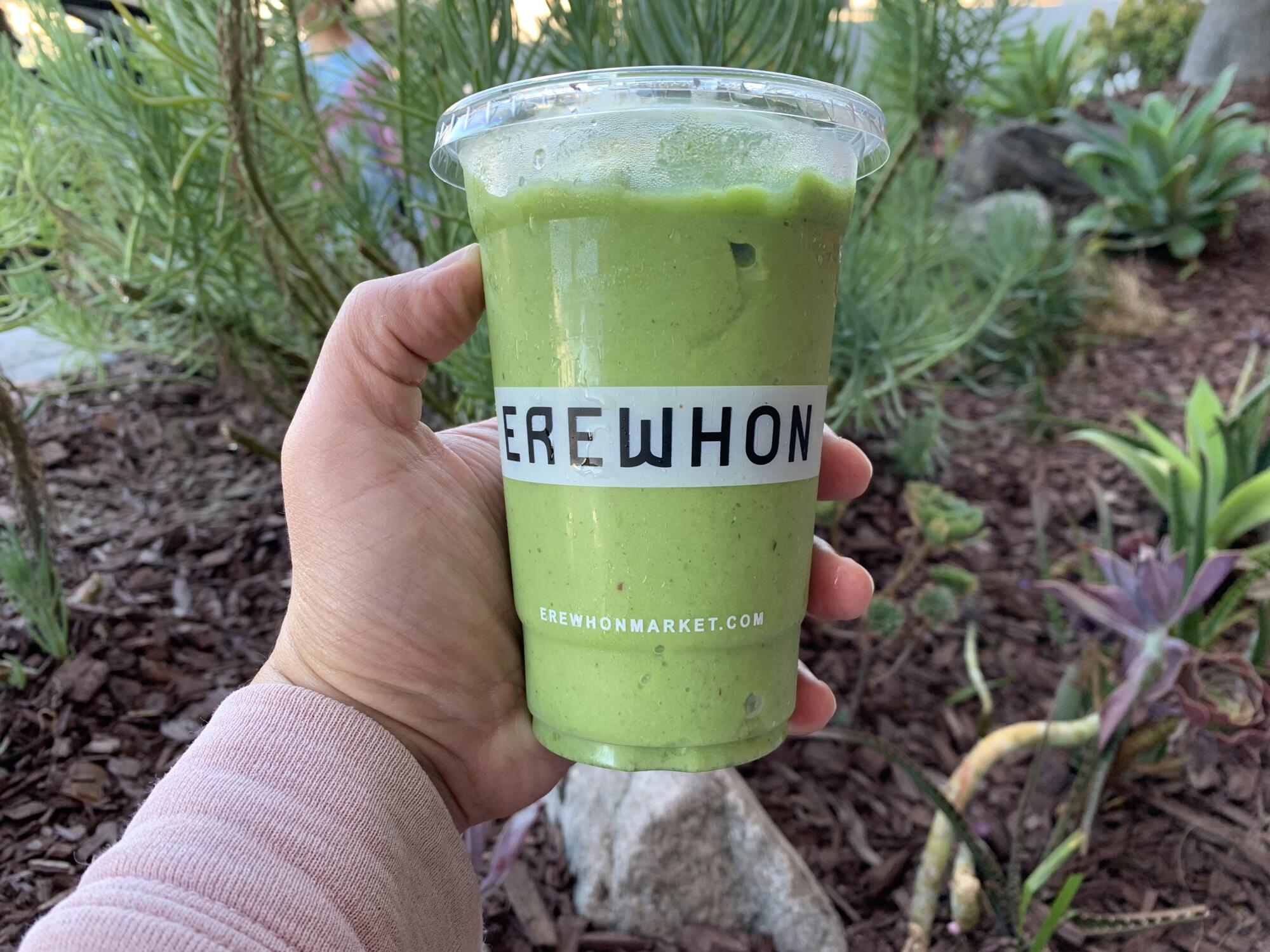 A hand holds a plastic cup containing a green smoothie.