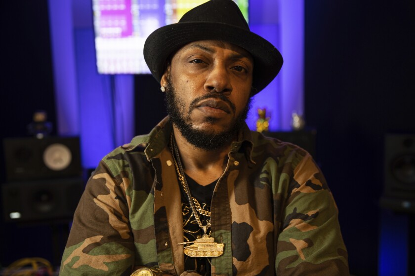 A beared man wearing a black hat, gold chains and camo jacket 