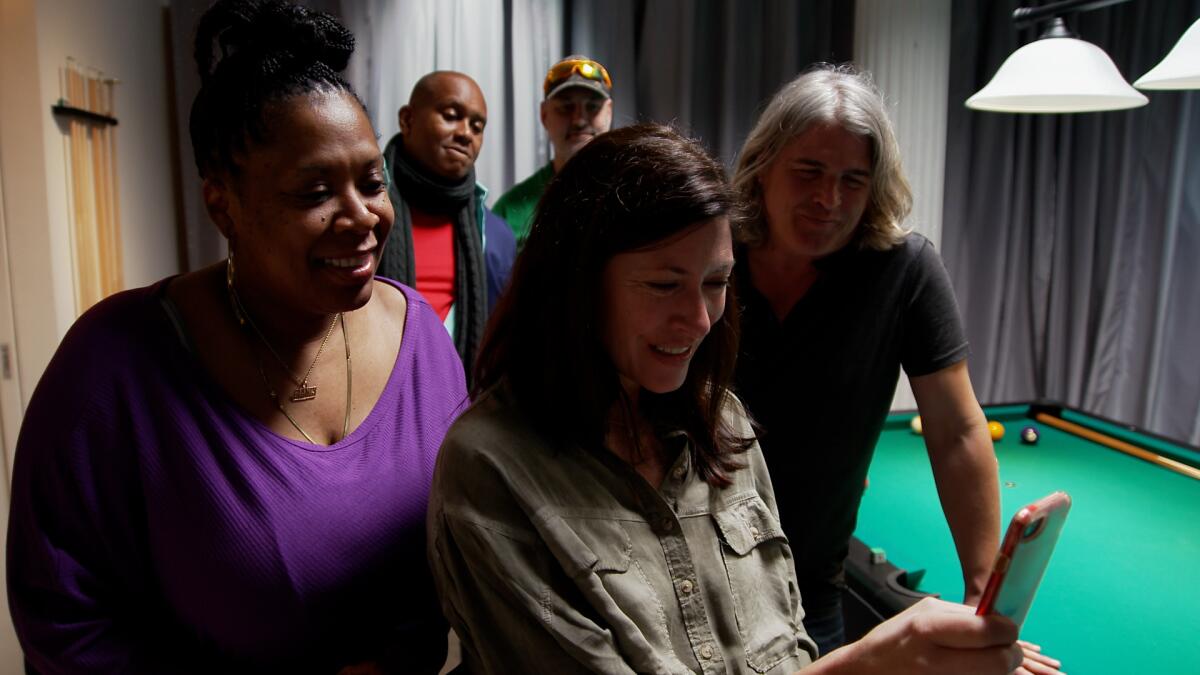 People stand in a room looking over the shoulder of a woman who's looking at her cellphone.