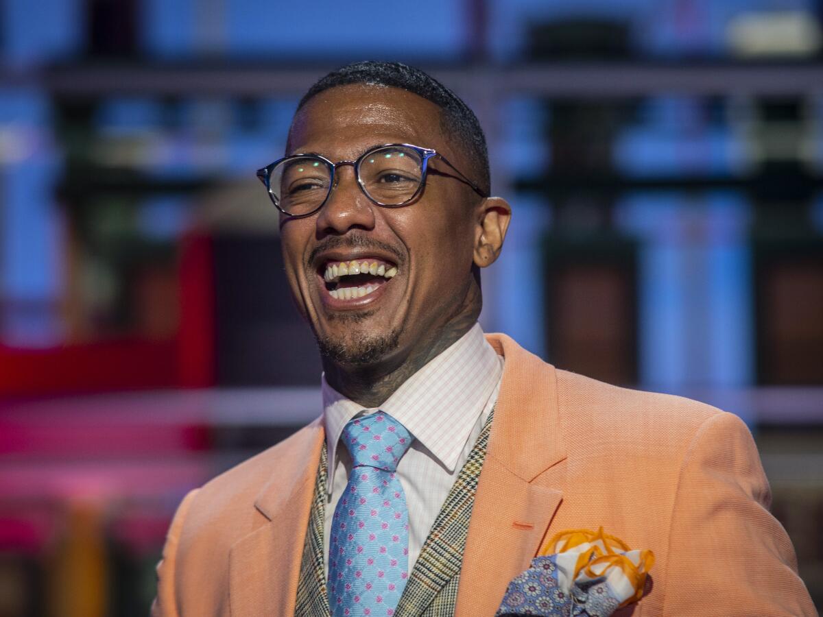Nick Cannon wears a burnt orange suit with a green vest, blue and red tie and a white shirt as he laughs 