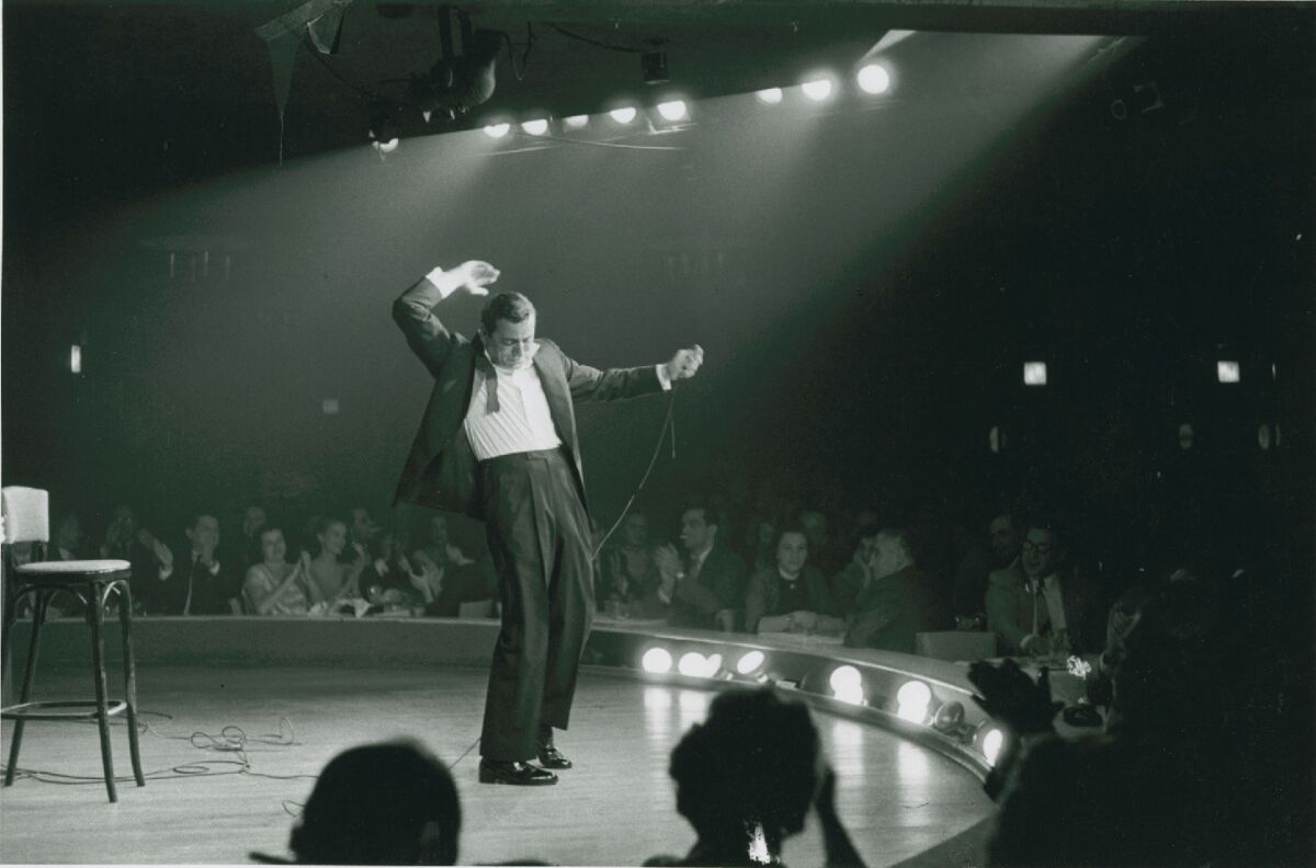 Tony Bennett dances and sings on a stage while people sit at tables near him.