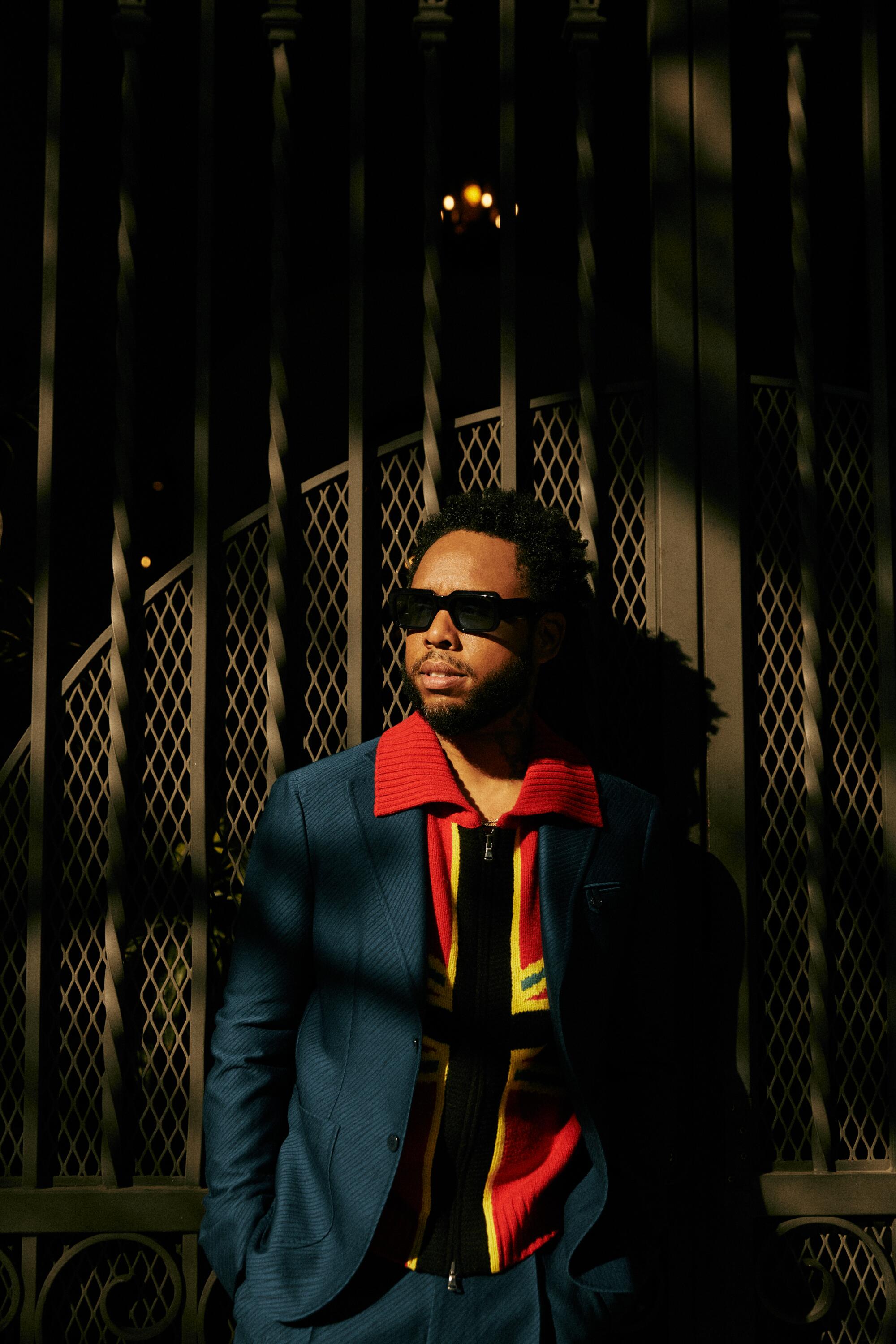 Terrace Martin stands against a wrought iron fence in a mix of shadow and sun.