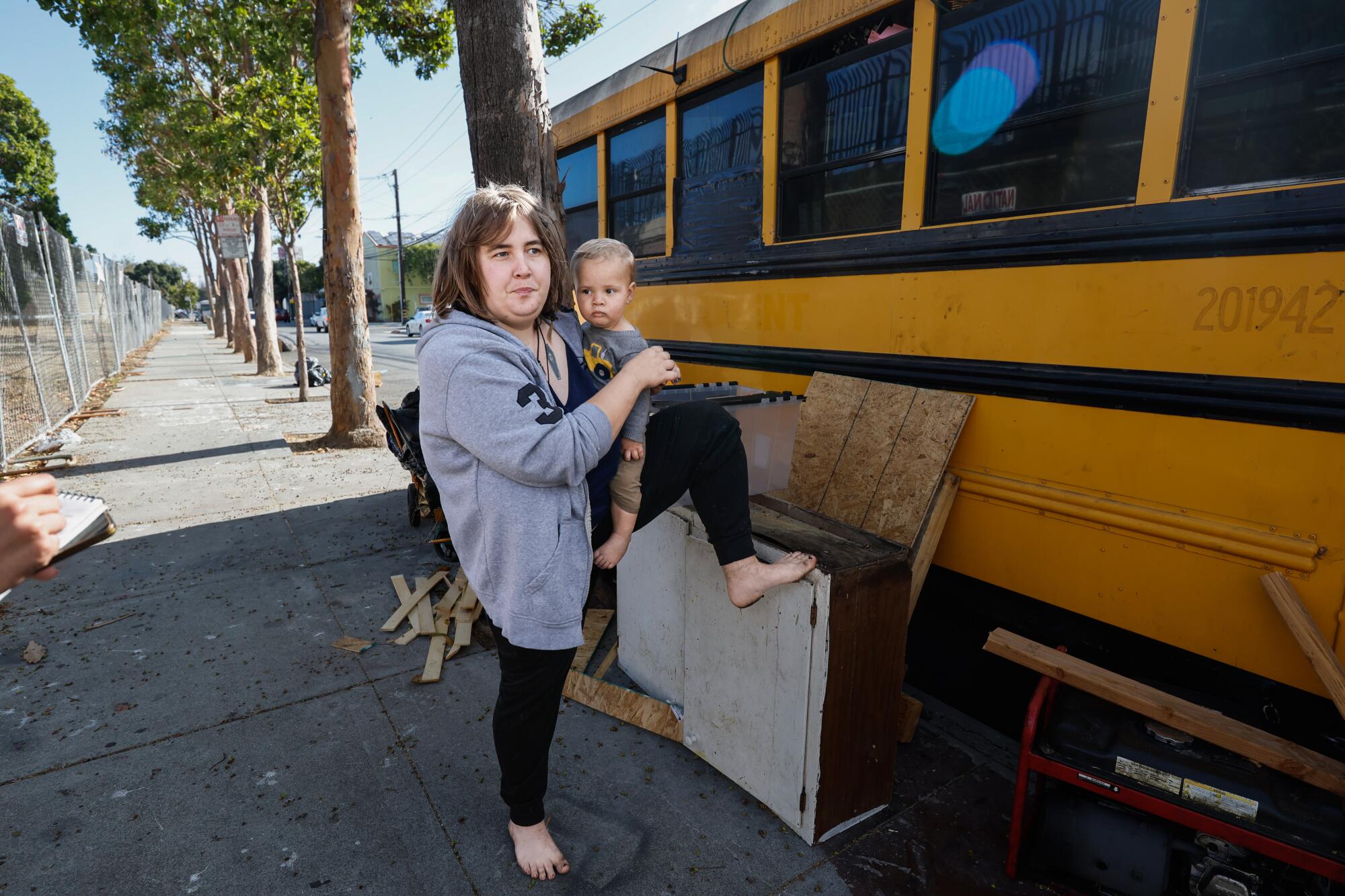 A woman holding an infant stands on a sidewalk beside a school bus