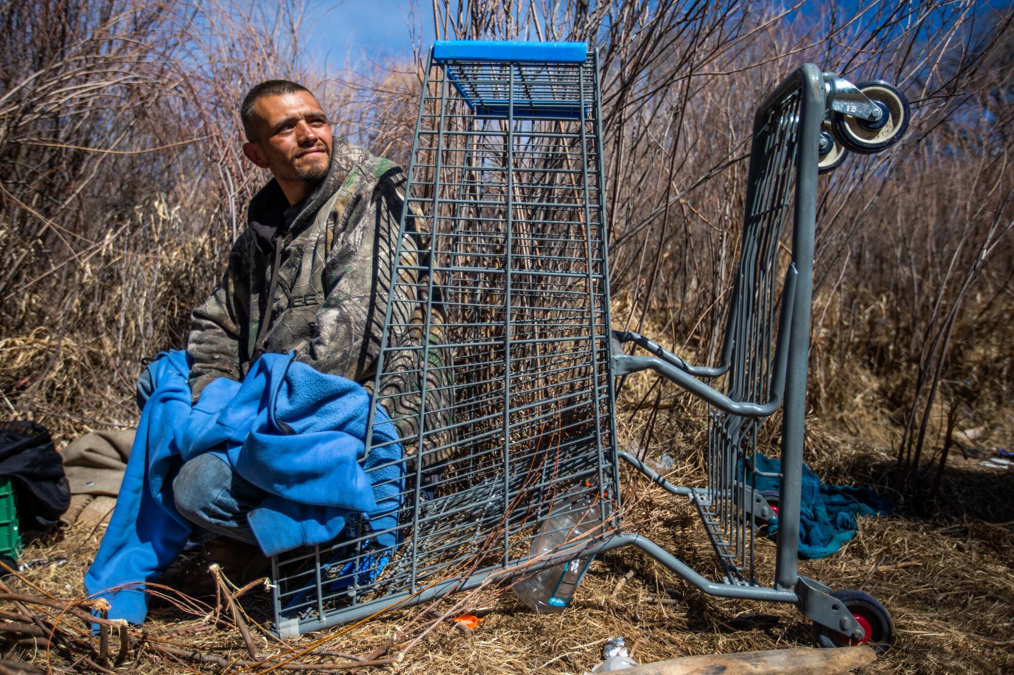 A man sits in an overturned shopping cart in a dry marsh amid little