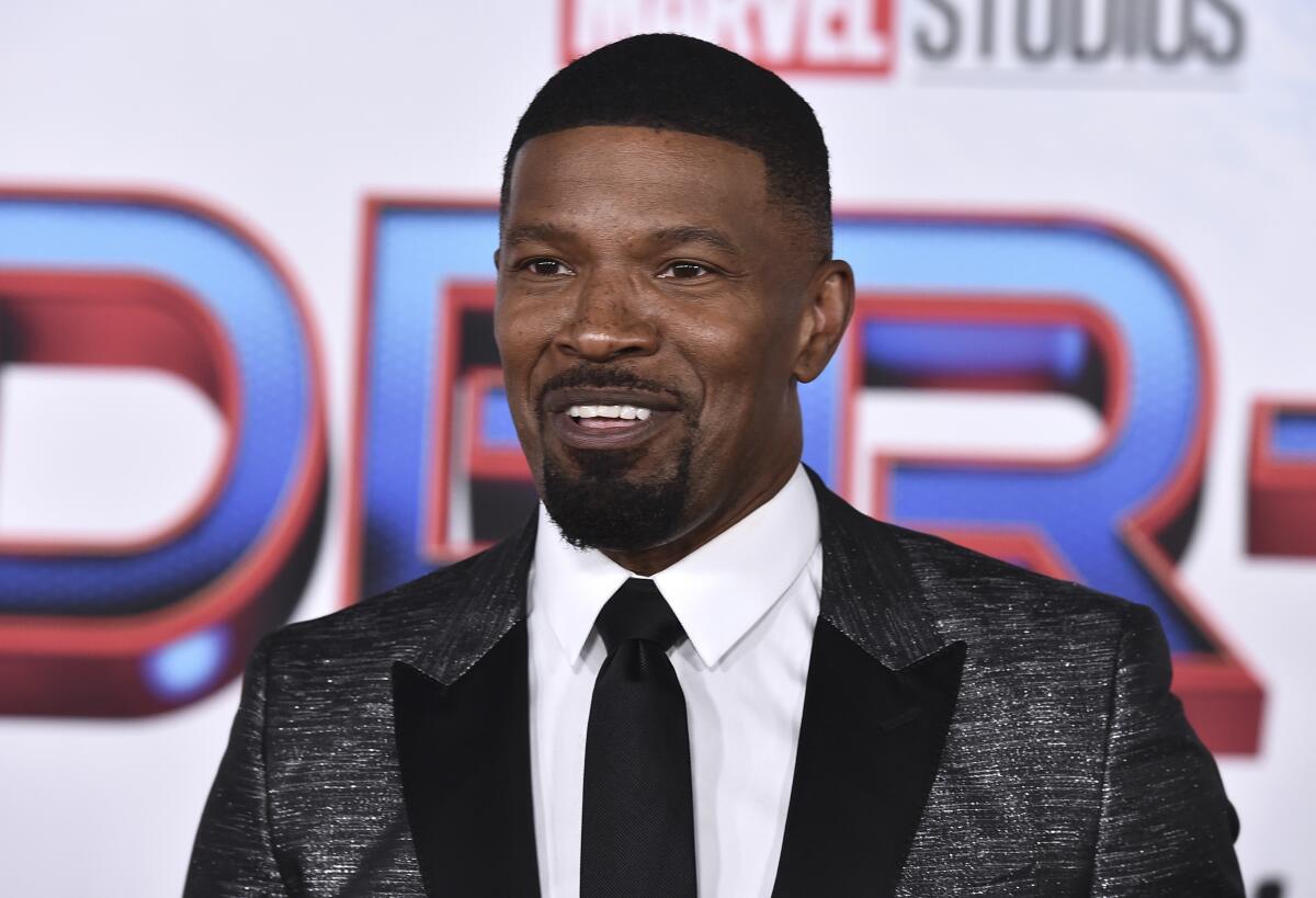 Jamie Foxx in a dark patterned suit and dark tie smiling at a red carpet event. 