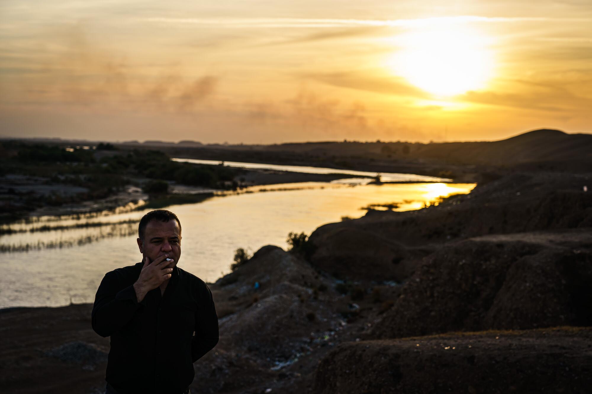 A man holding a cigarette stands in front of a river 