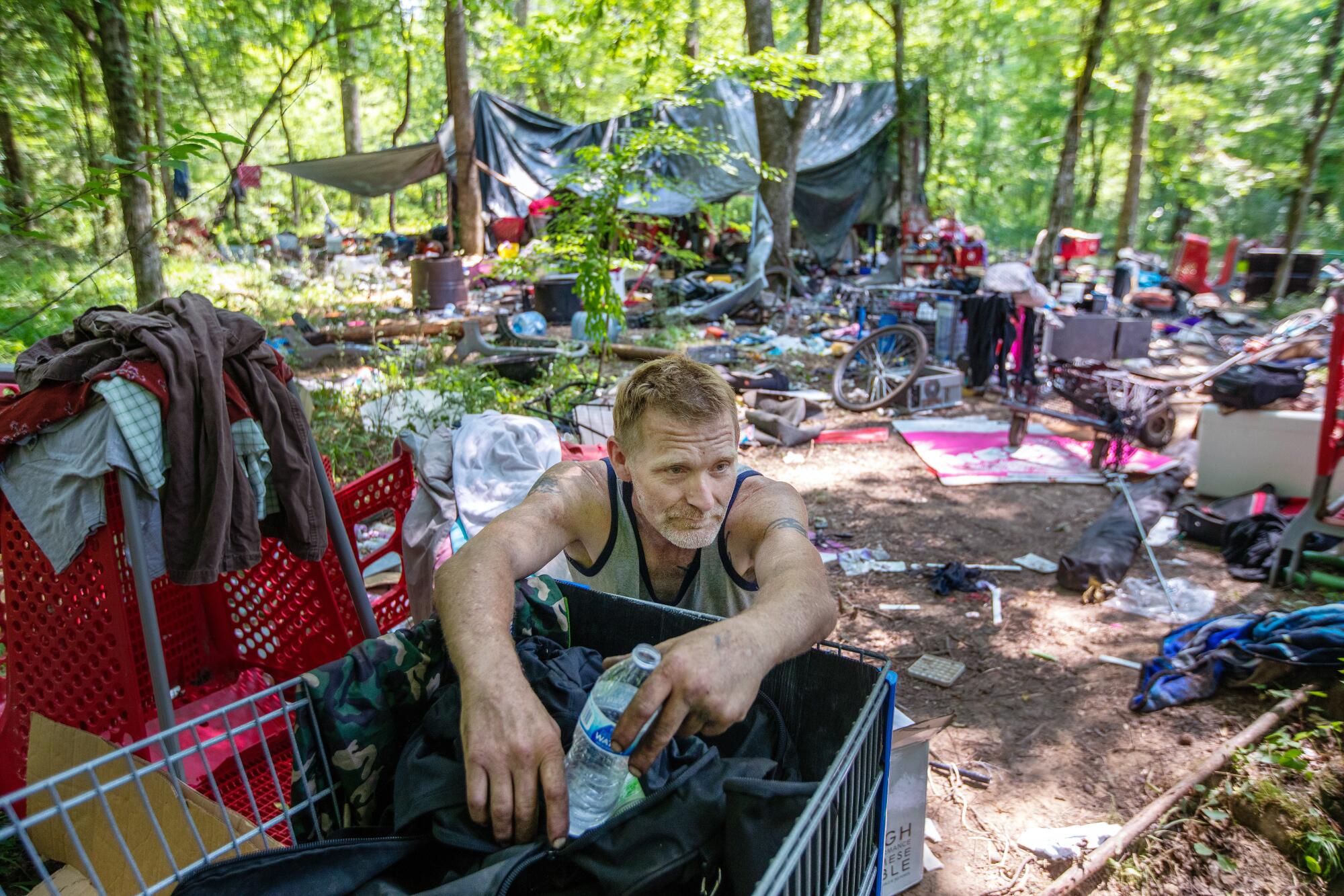 A man leans on a shopping cart in an outdoor encampment.