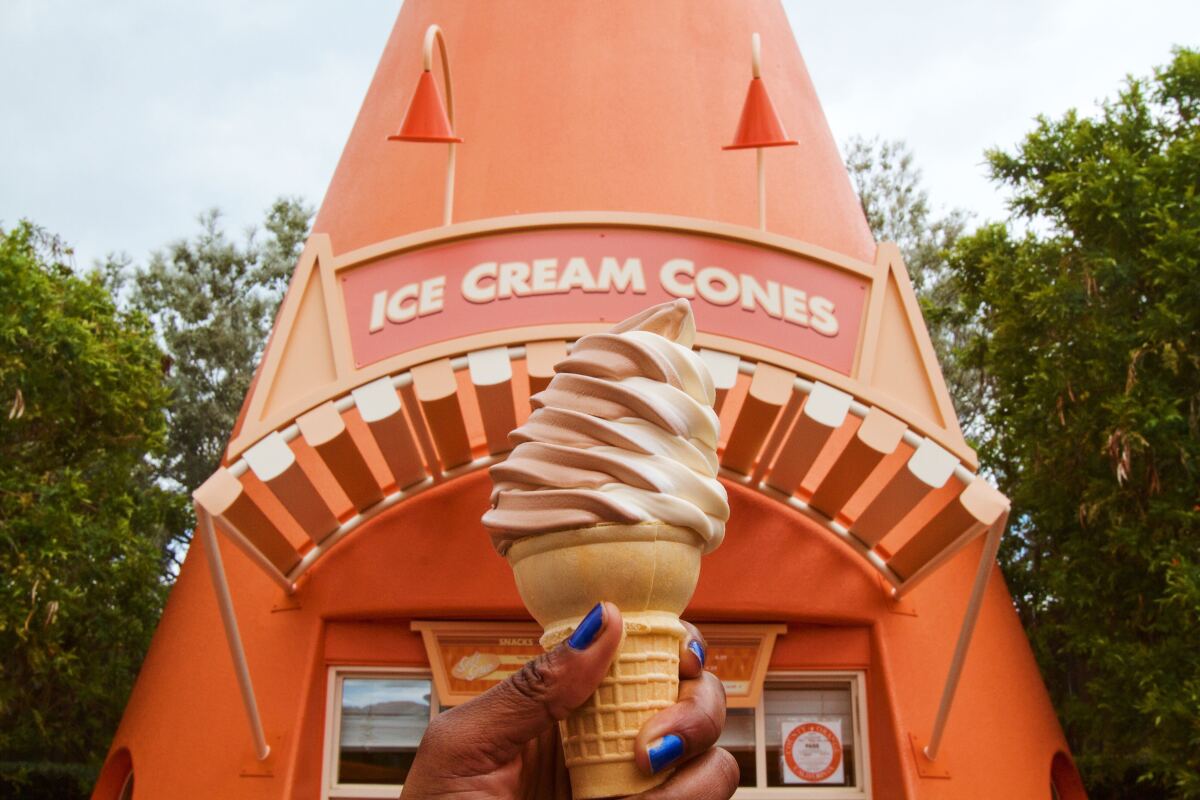 A hand holds up a chocolate and vanilla swirl soft-serve cone in front of an orange traffic-cone-shaped ice cream stand.