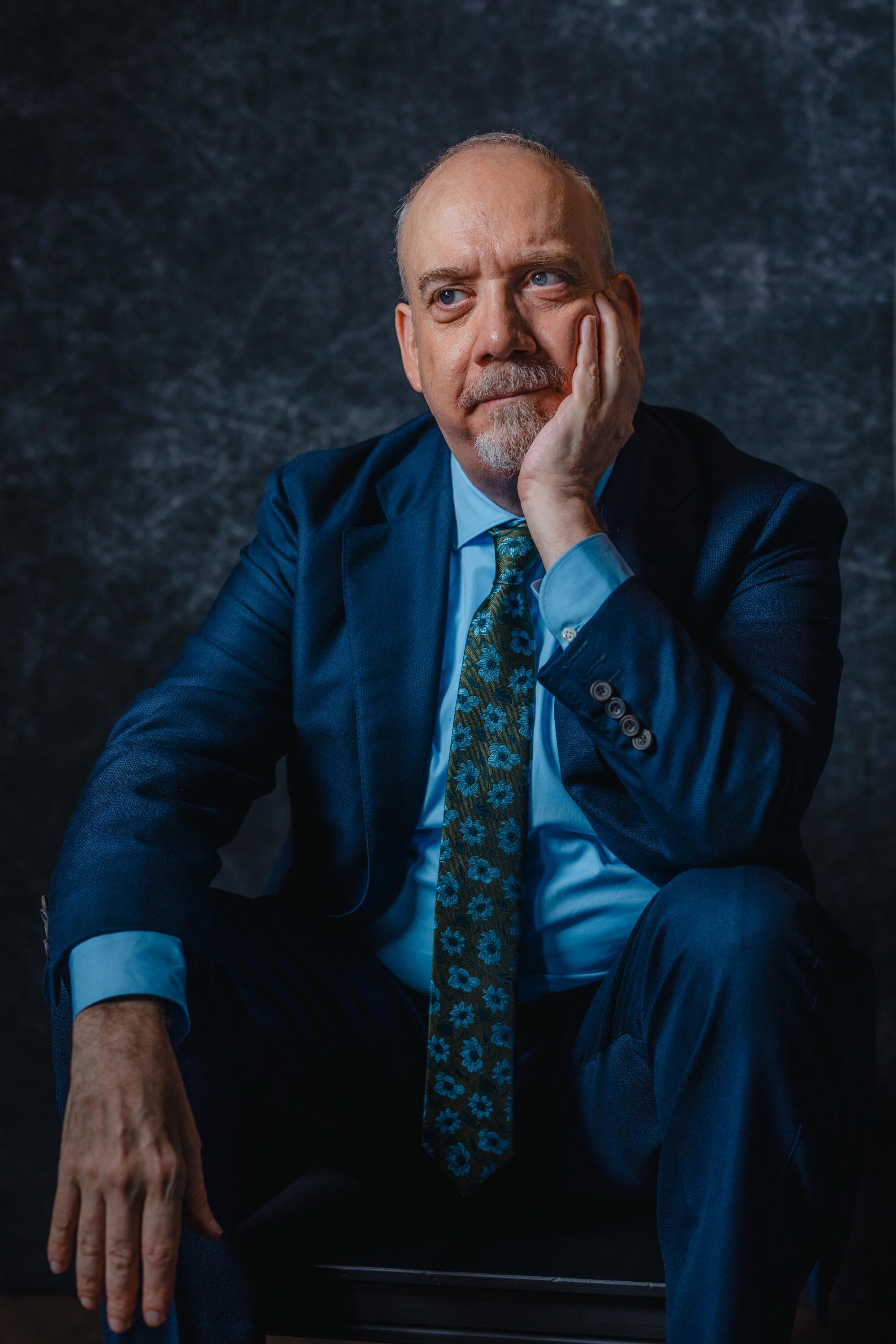 A grey-goateed man in a cobalt-blue suit sits wistfully, chin on his hand, before a dark blue background.