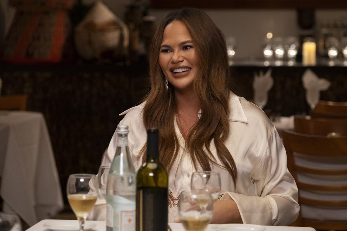 Chrissy Teigen in a white shirt sitting at a dining table.