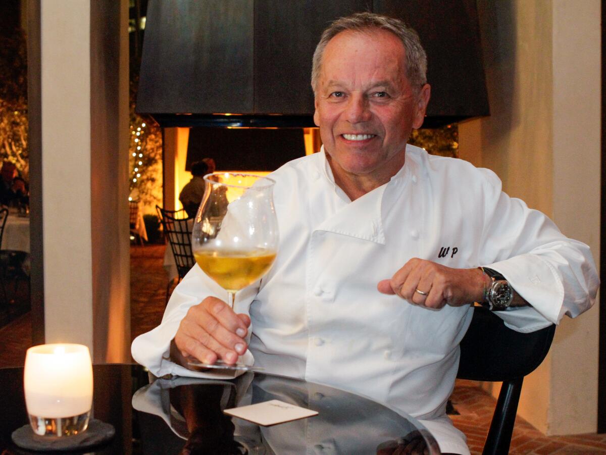 Wolfgang Puck wears a white chef's coat and holds a glass of wine at a table.