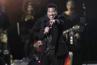 Lionel Richie is wearing a black suit and is pointing while performing on stage with a microphone held to his face
