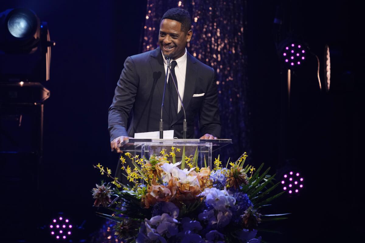 Blair Underwood wears a  black tuxedo as he speaks into a microphone at a podium onstage