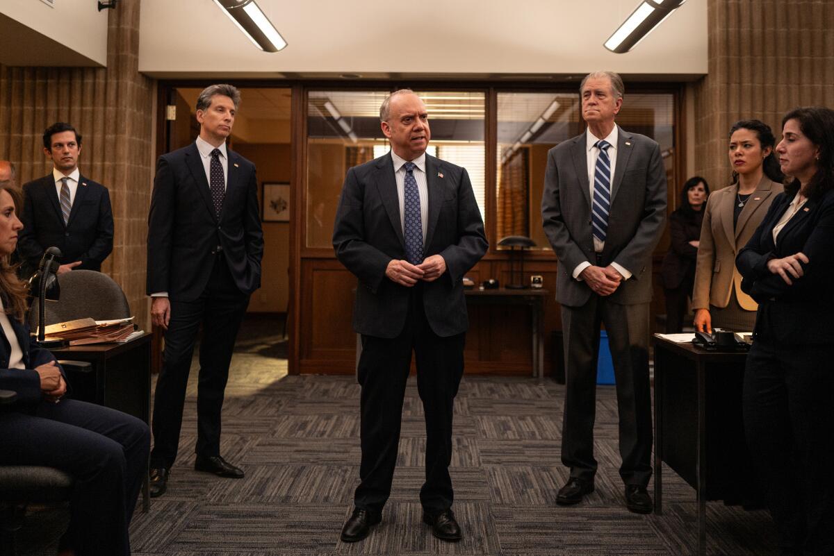 Three men in suits stand in the middle of an office as others look on.