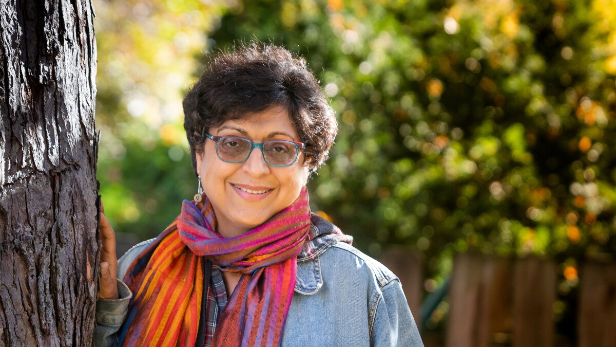 Thrity Umrigar stands smiling and leaning against a tree with her right hand resting on it