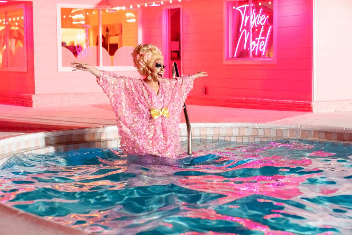 A drag queen stands in a round pool, arms spread wide