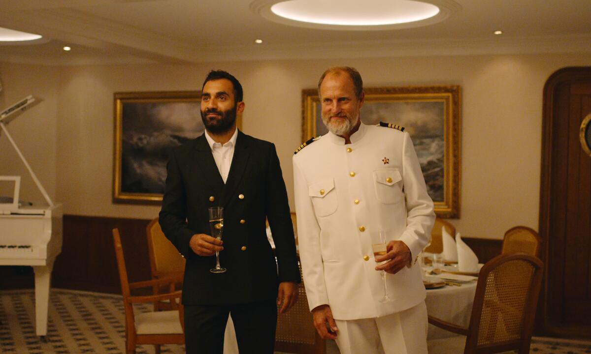 A man in a black suit stands next to a man in a white uniform in a scene from "Triangle of Sadness."