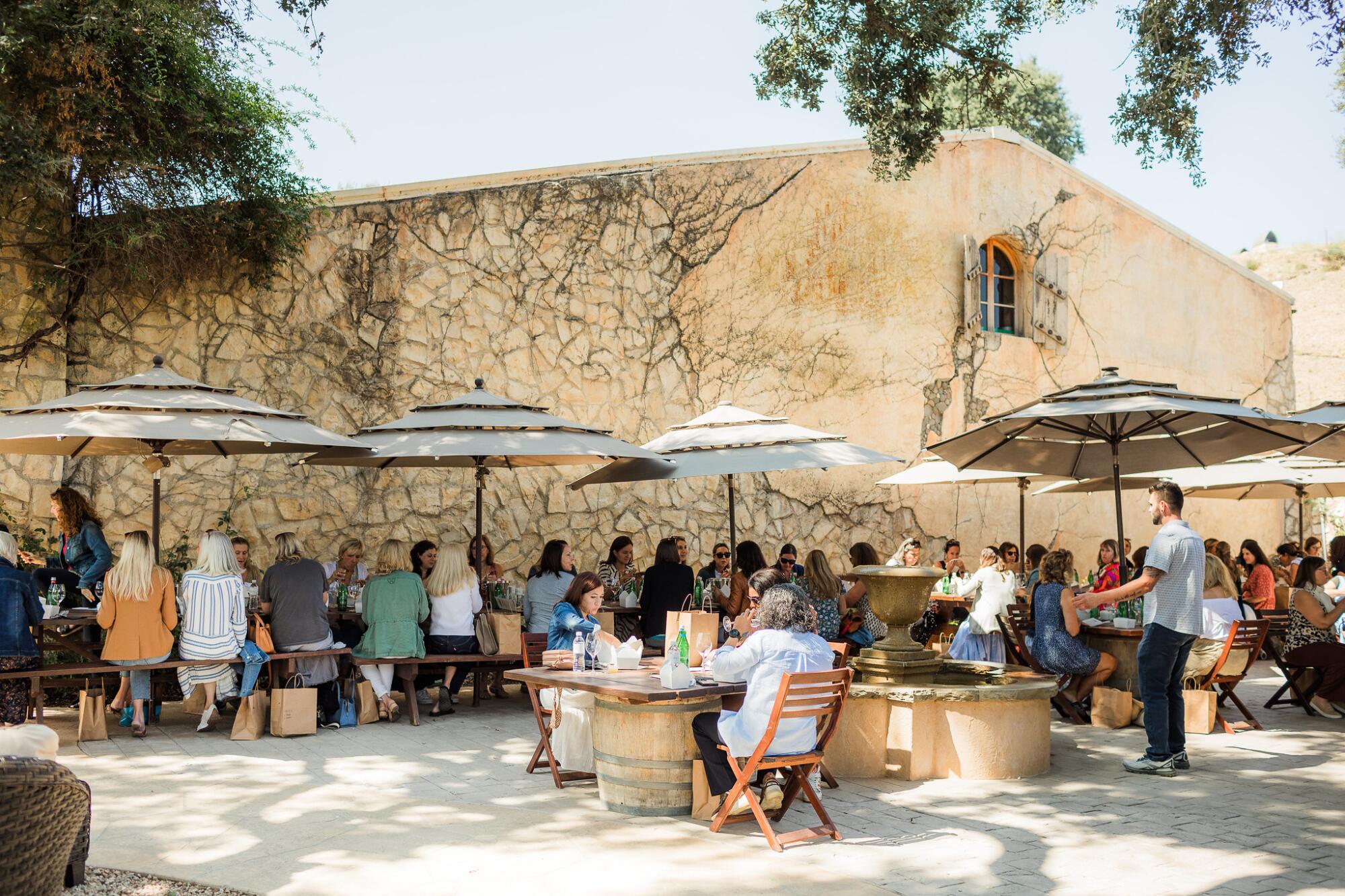 Women sit at long tables under umbrellas outside a low sandstone building.