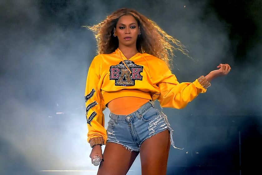A woman with long curly hair wearing a yellow hoodie and striking a pose on a stage