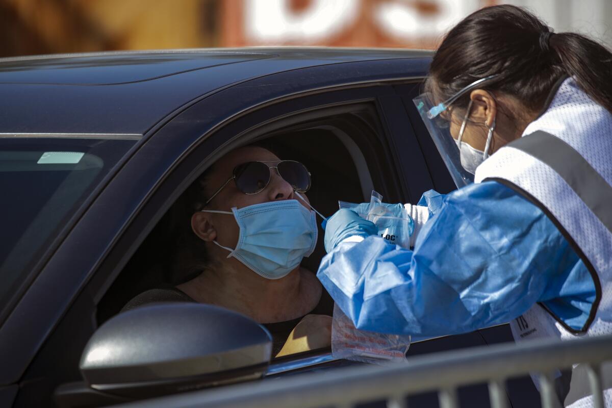 A woman in protective gear, gloves and goggles inserts a swab in the nose of a man as he sits behind the wheel of a car.