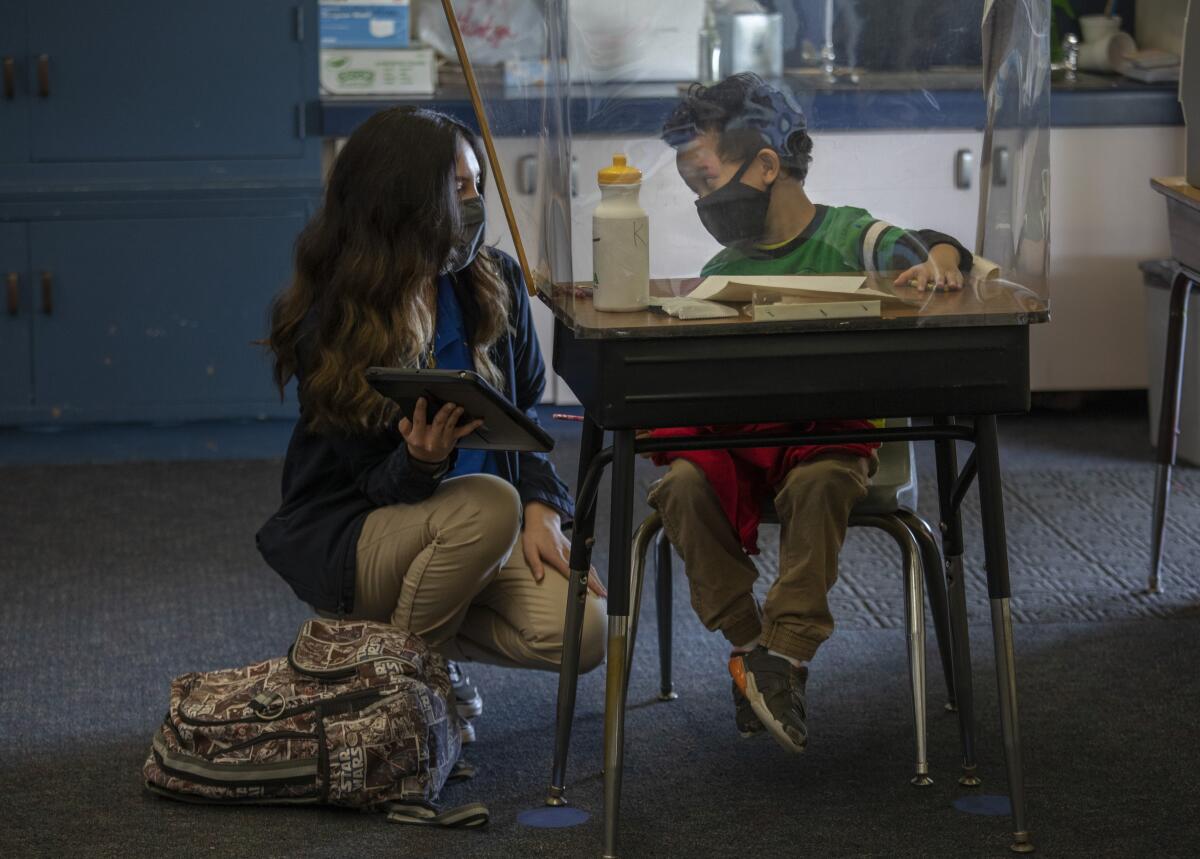 A transparent shield separates a classroom aide from a kindergartener.