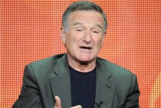 Robin Williams opens his mouth and gestures with his hands while sitting in front of an orange background