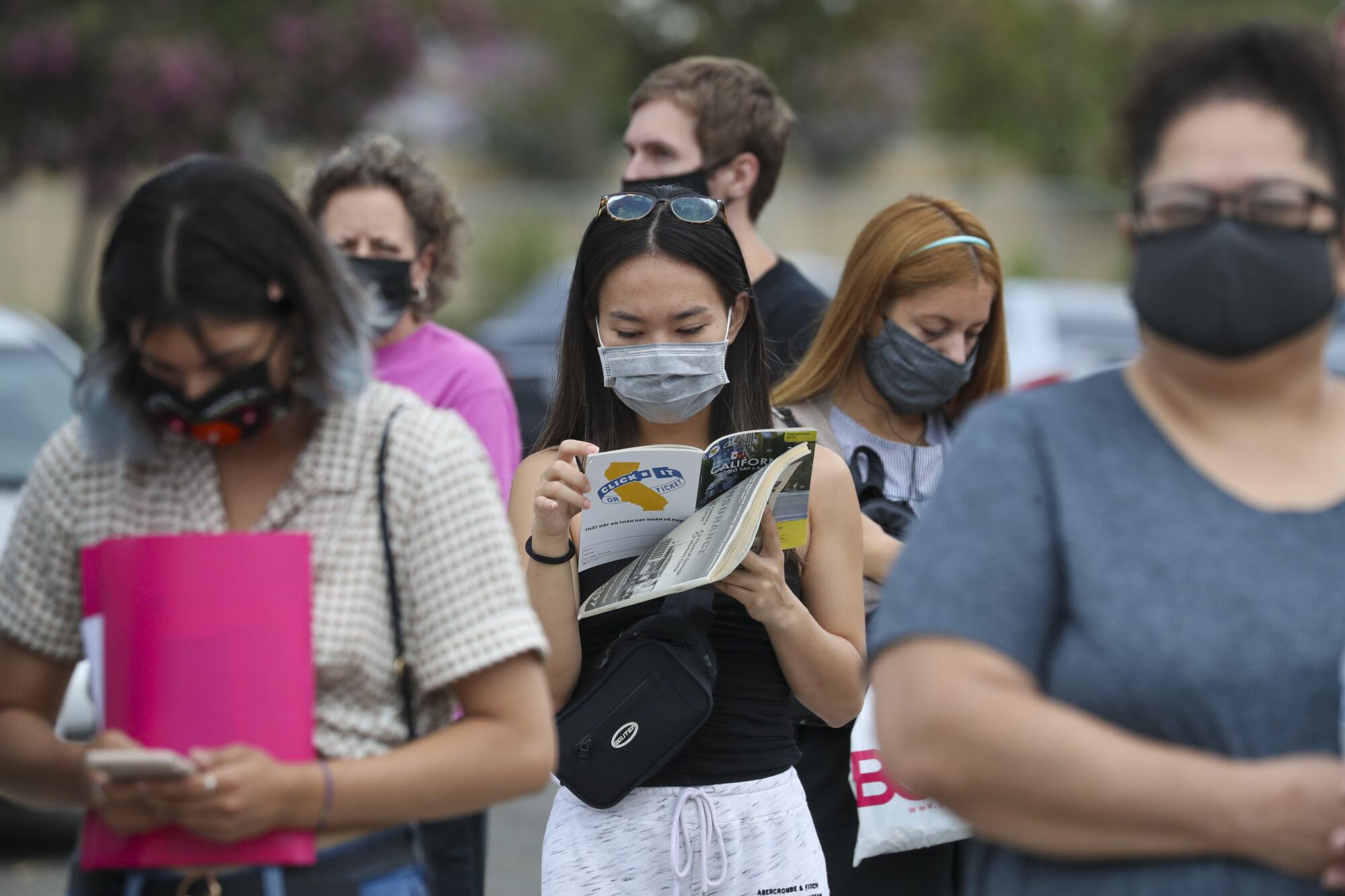 A woman wearing a mask looks down at a booklet while standing in line outside a DMV field office